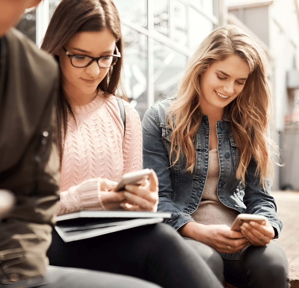 Students sitting outdoors on a university campus reading the Studo newsfeed on their smartphones